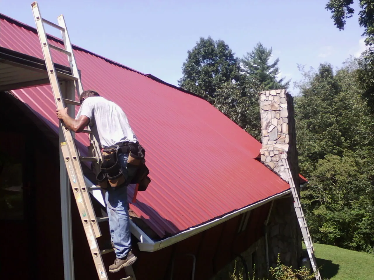 Roofer on a ladder beside a red metal roof and stone chimney — Soffit & Fascia Repair by Roofing Brothers in Mystic